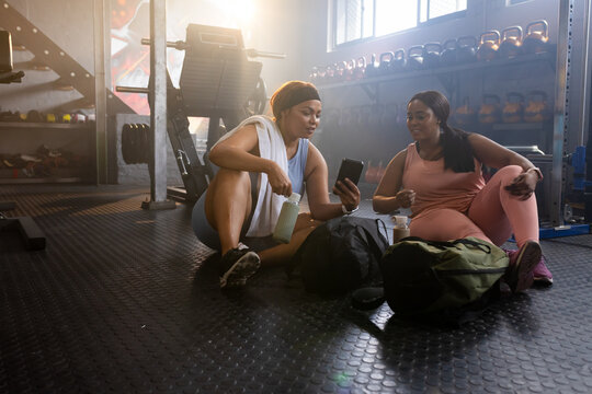 Female friends in gym clothes sitting on weight room floor, checking smartphone and holding bottle