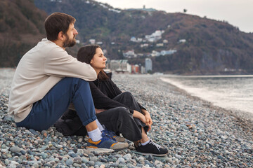 Couple sitting side by side on rocky beach, facing the sea with thoughtful expressions, enjoying peaceful moment of togetherness and mindfulness during calm evening