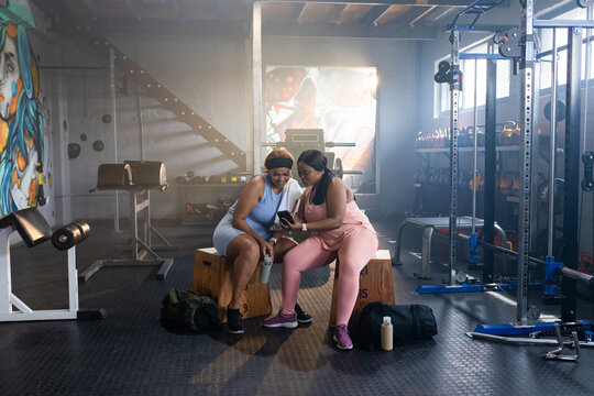 Diverse female friends in activewear sitting on wooden plyo boxes at gym, leaning in checking phone