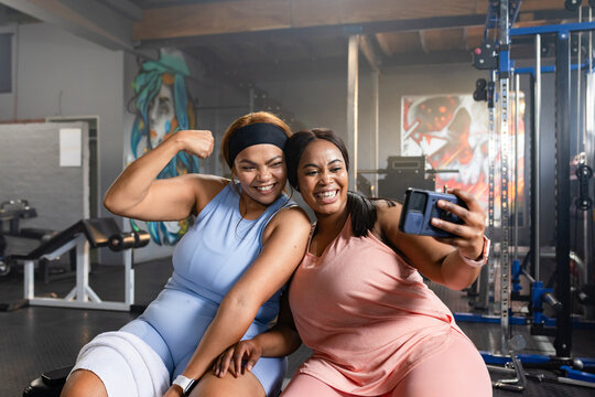 Diverse female friends sitting on bench in blue pink gear, flexing bicep taking selfie on phone
