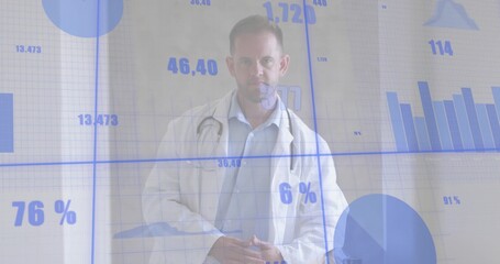 Standing clinician wearing white lab coat and stethoscope, facing camera at clinic with data charts