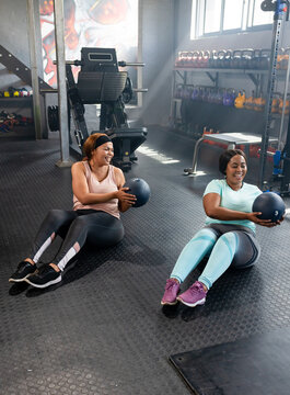 Diverse female friends in workout gear holding medicine balls and doing partner core work at gym