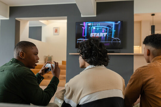 Diverse male friends sitting on sofa in living room holding controller and watching game screen