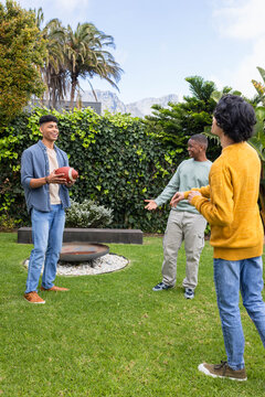 Diverse male friends standing in backyard holding football near fire pit in sneakers, copy space