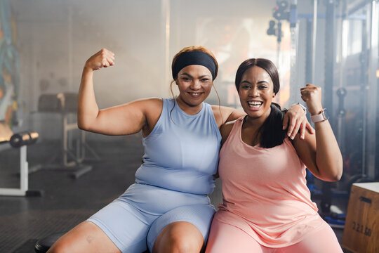 Diverse female friends posing and flexing in athletic wear on gym bench with dumbbells, earbuds