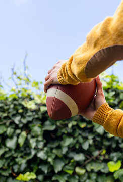 Hands and forearms are gripping American football in backyard, showing mustard sweater and hedge