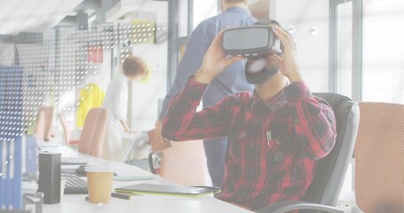 Holding VR headset, man in red plaid shirt testing virtual app at open-plan office, with laptop