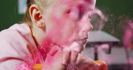 Leaning child in pink sweater, folded hands on orange chair back in studio, pink dust drifting