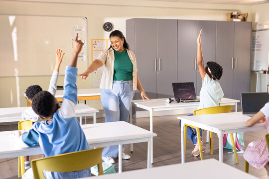 African American teacher in jeans pointing to raised hands engaging youth with laptops in class