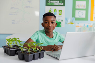 African male child sitting at classroom table folding hands beside silver laptop and seedling trays