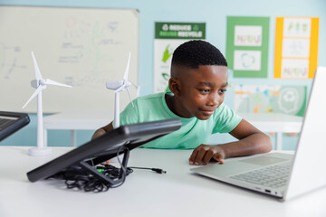 African male youth testing laptop, wind turbines and black charger in STEM lab wearing green tee