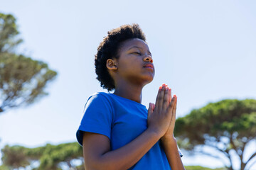 African American female teen standing under clear sky wearing blue tee, meditating by canopy trees