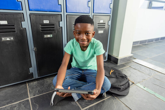 African American child sitting cross-legged on tiled hallway holding tablet and backpack by lockers