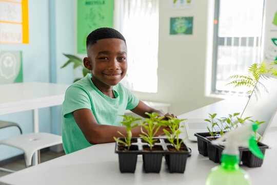 African American school-age boy smiling and tending seedling trays at classroom table using laptop