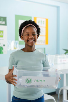 African American female youth smiling and holding clear bin labeled PAPER in school recycling area