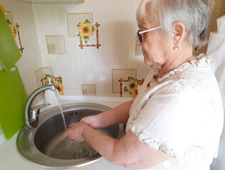 An elderly woman washes her hands with soap under running water from a kitchen sink faucet.
