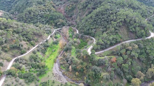 An aerial drone shot ascending and slowly pulling back over a winding mountain road in the Genal Valley, Andalusia, Spain. The footage reveals a scenic winter landscape with green and ochre tones