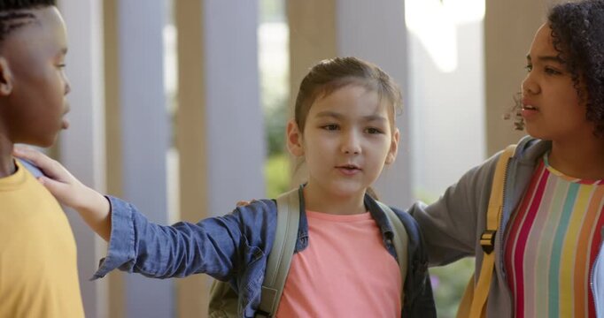 Diverse kids at school porch calming after spat, center girl placing hands on shoulder with packs