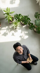 Young man sitting on chair holding notebook, surrounded by green indoor plants, soft natural light, relaxed and thoughtful mood, modern home interior, top view
