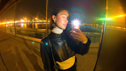 Young woman taking selfie at night with city lights reflecting on water, wearing black leather jacket and white turtleneck, standing outdoors by waterfront, vibrant and confident mood
