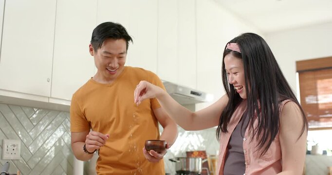 Asian couple in kitchen, man offering bowl and spoon, woman drizzling syrup, twirling while cooking