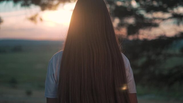 Girl with longhair enjoys peaceful sunset. Backview in field shows silence of nature. Landscape spreads in calm solitude. Girl reflects on nature. Sunset lights girl and longhair in reflection.