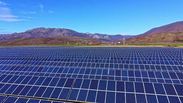 Rows of solar panels are set up in an open field at a renewable energy farm. Mountains rise in the background under bright blue skies. A wind turbine is visible in the distance.
