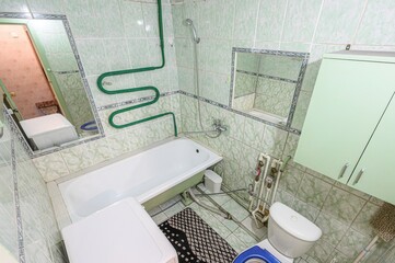 A compact bathroom with a bathtub, toilet, wall-mounted cabinet, green heated towel rail, tiled walls, two mirrors, exposed plumbing pipes, bath mat, and storage unit.