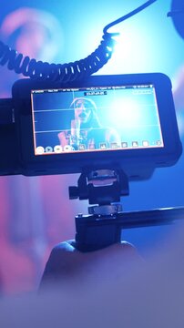 Behind the scenes look at a film set. Videographer's hand holds a camera with an external monitor, framing a singer performing at a concert. Vertical footage
