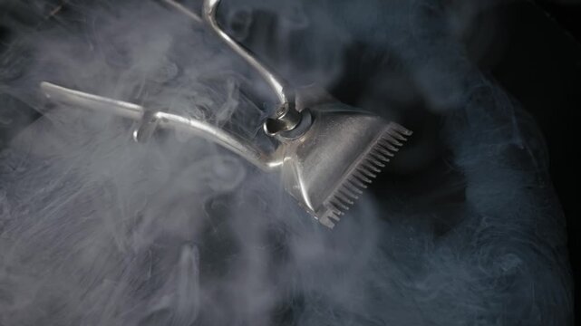 Vintage manual hair clipper on a dark surface is enveloped by thick white smoke. The mysterious vapor swirls around the metallic barber tool in a dramatic slow motion shot.