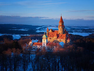 Panoramic aerial view of Bouzov Castle rising above snowy forest and rolling Czech countryside at golden hour