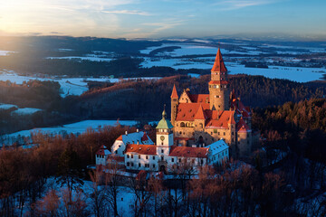 Panoramic aerial view of Bouzov Castle rising above snowy forest and rolling Czech countryside at golden hour
