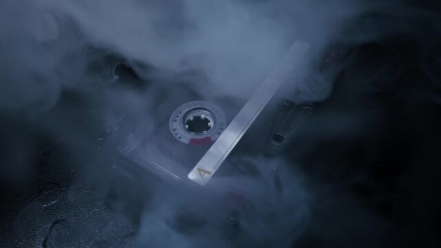 Vintage audio cassette tape with an 'A' label lies on a dark surface. Mysterious smoke drifts across the tape in a dark, atmospheric setting.