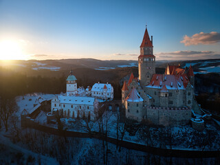 Panoramic aerial view of Bouzov Castle rising above snowy forest and rolling Czech countryside at golden hour