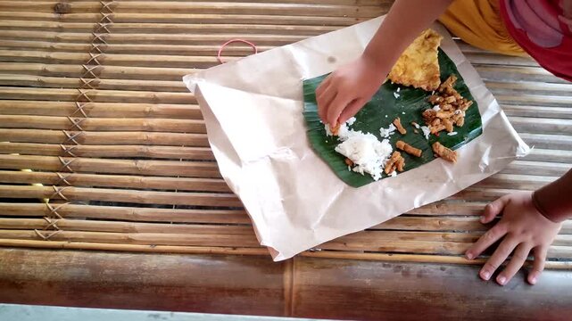 Young child eating a traditional Indonesian meal (Nasi Bungkus) with their hands. The meal consists of white rice, fried tempeh, and an omelette served on a banana leaf and brown paper