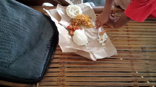 A young child sits while enjoying a typical Indonesian food known as &ldquo;Nasi Bungkus&rdquo;. The meal, served on brown wrapping paper, includes white rice, stir-fried noodles, and a large white cracker