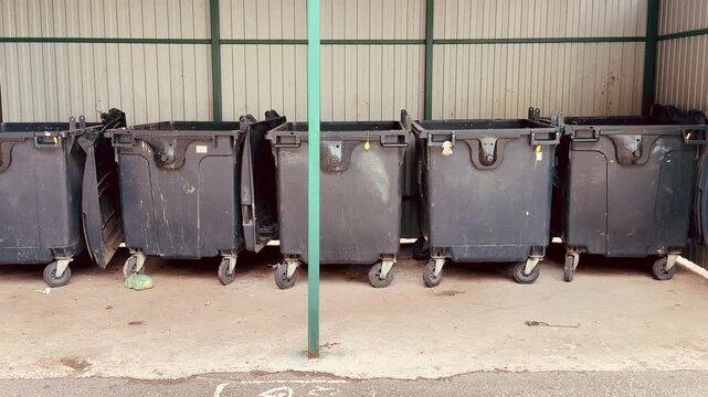 black dumpsters on concrete alley, closeup of worn caster wheels, scuffed lids and chipped paint, scattered debris and small puddles on pavement, industrial fence background,