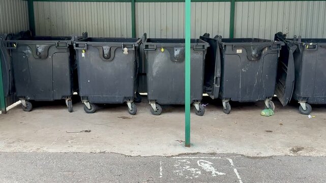 black dumpsters on concrete alley, closeup of worn caster wheels, scuffed lids and chipped paint, scattered debris and small puddles on pavement, industrial fence background,