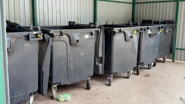 black dumpsters on concrete alley, closeup of worn caster wheels, scuffed lids and chipped paint, scattered debris and small puddles on pavement, industrial fence background,