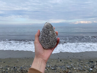 Hand holding smooth stone by ocean on cloudy day with waves and rocks on shore