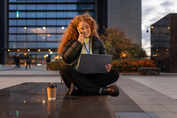 Redhead woman working outdoors using laptop and talking on phone at a modern city building