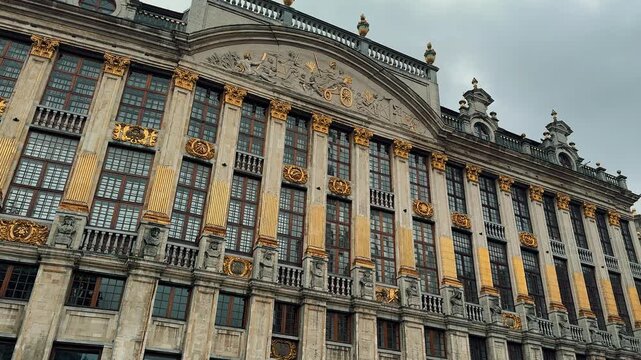 Slow pan shot across the magnificent, gilded facade of a historic guildhall on the Grand Place in Brussels, Belgium, showcasing its ornate architectural details under an overcast sky.

