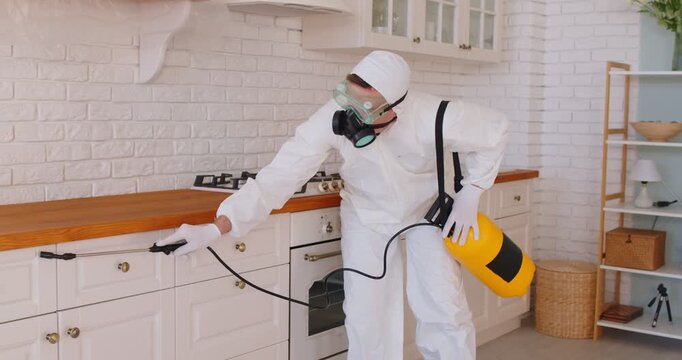 Pest control service disinfestation exterminator and maintenance technician in hazmat uniform uses household insecticide to sanitize residential home kitchen from domestic insect contamination