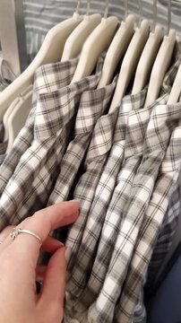 Woman's hand with a ring browses a rack of grey and white plaid flannel shirts in a store. Close-up shot follows her fingers as they touch the soft fabric of the clothing.