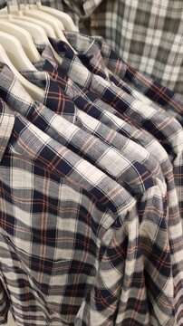 Plaid flannel shirts hang in a row on clothes hangers inside a retail store. The camera pans up, revealing the soft fabric texture and checkered pattern.