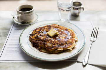 Pancakes with warm maple syrup and butter on a white plate in a restaurant setting.