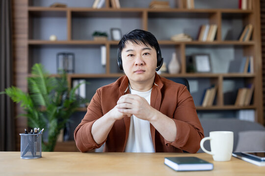 Asian man sitting at desk wearing a headset, participating in an online meeting or webinar, working remotely from a home office with a bookshelf in the background - Powered by Adobe