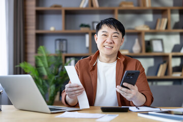 Asian man smiling and holding a receipt and smartphone while sitting at a desk with a laptop and calculator, organizing personal finances and taxes at his home office