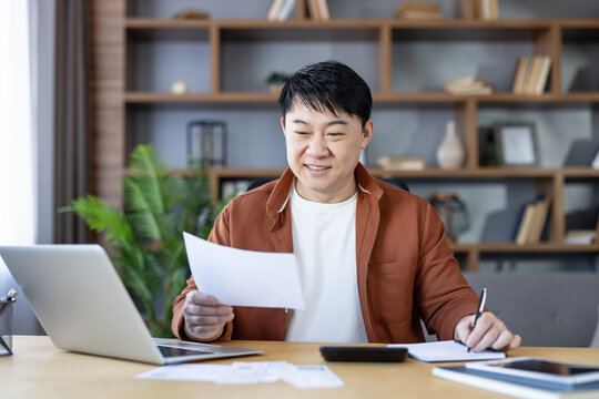 Smiling asian man analyzing documents and making notes at his laptop in a home office, proactively planning personal finances, expenses, and investment strategies