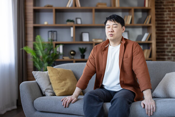Asian man sitting relaxed on a comfortable sofa with closed eyes, engaging in a mindful breathing exercise or meditation for stress relief and mental well-being in his living room